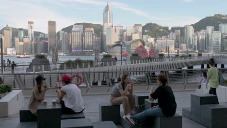 Man-Sieht-Leute,-Die-Draußen-An-Der-Victoria-Waterfront-Sitzen-Und-Trinken,-Während-Die-Ikonische-Hölzerne-Dschunke-Mit-Den-Roten-Segeln,-Bekannt-Als-Aqua-Luna,-über-Den-Hafen-Und-Die-Skyline-Von-Hongkong-Segelt