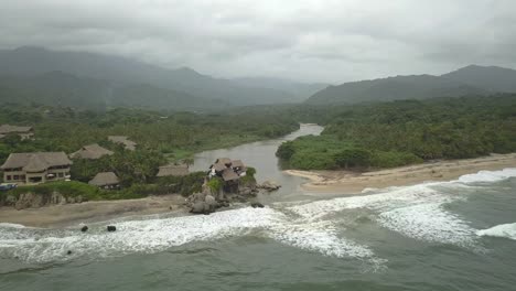 Wunderschöne-Drohnenaufnahme-Eines-Strandes-Und-Eines-Dschungelflusses-Im-Tayrona-Nationalpark-In-Kolumbien-Mit-Bergen-Im-Hintergrund