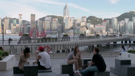 Man-Sieht-Leute,-Die-Draußen-An-Der-Victoria-Waterfront-Sitzen-Und-Trinken,-Während-Das-Ikonische-Hölzerne-Dschunkenboot-Mit-Den-Roten-Segeln,-Bekannt-Als-Aqua-Luna,-über-Den-Hafen-Und-Die-Skyline-Von-Hongkong-Segelt