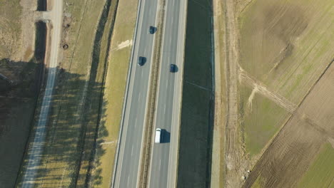 La-Luz-Del-Sol-Baila-A-Través-De-Una-Carretera,-Proyectando-Largas-Sombras-Desde-Una-Fila-De-Vehículos-En-Tránsito,-Frente-A-Un-Paisaje-Rural