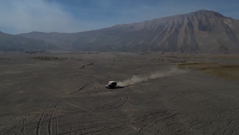 Siguiendo-Un-Coche-Con-Un-Dron-A-Través-De-Los-Campos-De-Lava-De-Bromo-Al-Amanecer,-Con-Llamativas-Huellas-De-Neumáticos-De-Arena