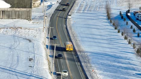 Yellow-school-bus-driving-on-road-in-rural-USA-surrounded-by-snow-covered-landscape-in-winter