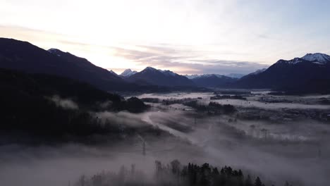 Drone-fly-above-misty-pine-forest-with-Aerial-Drone-View-over-cityscape-in-Frastanz,-Austria,-Sunrise-above-foggy-winter-mountain-landscape