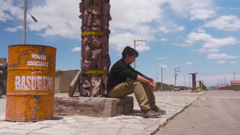 Einsamer-Mann-Sitzt-An-Einer-Felssäule-Neben-Einer-Mülltonne-Auf-Einer-Leeren-Straße-In-Bolivien