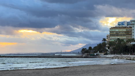 Marbella-beach-on-a-windy-cloudy-day-with-palm-trees,-apartments-and-mountains-in-Spain,-interesting-sunset-sky,-4K-shot