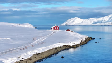 Red-house-along-fjord-in-Iceland,-in-a-snow-covered-landscape,-aerial-pan