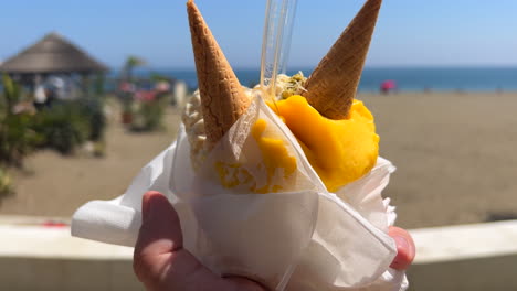 Halten-Sie-An-Einem-Sonnigen-Tag-Einen-Pappbecher-Mit-Mango-Eissorbet-Und-Waffeltüten-Am-Strand-In-Marbella-Estepona,-Spanien,-Süßes-Sommerdessert,-Urlaub,-4K-Aufnahme