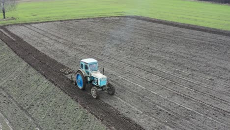 Aerial-view-of-old-tractor-equipment-cultivate-farmland,-emit-blue-exhaust-gas