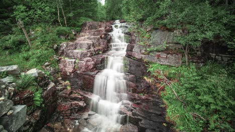 A-small-waterfall,-surrounded-by-greenery-in-the-summer-forest,-cascades-over-the-dark-stones