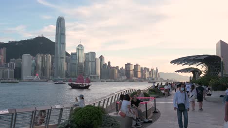 Die-Menschen-Genießen-Ihren-Nachmittag-An-Der-Victoria-Waterfront,-Während-Die-Ikonische-Hölzerne-Dschunke-Mit-Den-Roten-Segeln,-Bekannt-Als-Aqua-Luna,-über-Den-Hafen-Und-Die-Skyline-Von-Hongkong-Segelt,-Während-Die-Sonne-Langsam-Untergeht