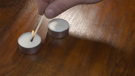 Right-hand-uses-burning-wooden-match-to-light-two-tea-candles-on-table