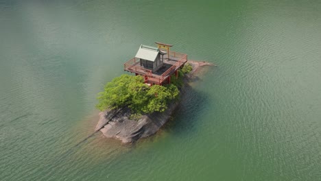 Aerial-drone-fly-above-temple-alone-in-the-middle-of-the-water-of-Kyoto-japan-water-landscape,-religious-orange-Shinto-entrance-with-Tori-doors-and-wooden-architecture-at-blue-lake
