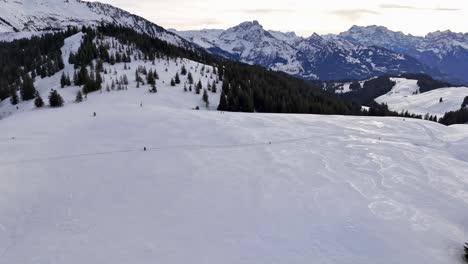 Weite-Schneelandschaft-In-Der-Schweiz-Mit-Skifahrern-In-Der-Ferne-Und-Bergkulisse,-Luftaufnahme
