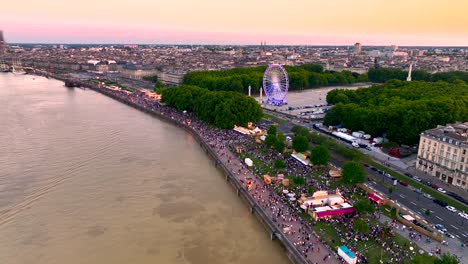 Wine-fair-at-Garonne-river-shore-with-Ferris-wheel-and-crowds-at-sundown,-Aerial-tilt-down-rising-shot