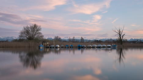 Calm-sunset-on-a-lake-with-snowy-mountains-in-the-background-and-boats-on-the-water