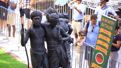 39th-Infantry-Battalion-banner-next-to-the-bronze-statue-of-the-South-West-Pacific-Campaign-with-crowds-on-the-pathway-of-Anzac-square,-honouring-those-who-served-during-wartime,-close-up-shot