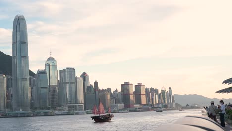 Vista-Del-Icónico-Barco-Chatarra-De-Vela-Roja-De-Madera-Conocido-Como-Aqua-Luna-Navegando-Por-El-Puerto-De-Victoria-Y-El-Horizonte-De-Hong-Kong-Mientras-La-Puesta-De-Sol-Comienza-A-Asentarse