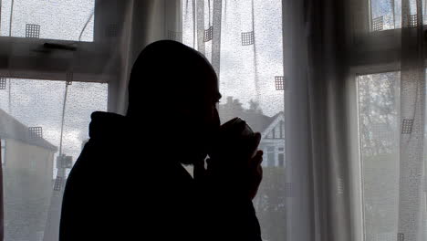 Standing-beside-the-window,-silhouette-of-bald-man-savors-his-coffee-while-gazing-at-the-daytime-scene-outside,-exemplifying-the-essence-of-routine-household-moments
