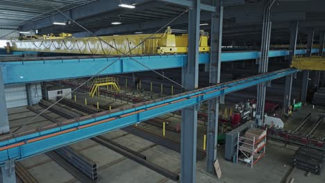 Aerial-descend-inside-of-steel-pipe-beam-warehouse-establishes-yellow-overhead-bridge-crane