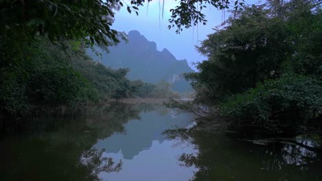 Blick-Auf-Die-Dichte-Baumvegetation,-Die-Sich-An-Einem-Bewölkten-Tag-In-Einem-Kleinen-See-Mit-Der-Silhouette-Zerklüfteter-Berge-Im-Hintergrund-In-Ninh-Binh,-Vietnam-Widerspiegelt