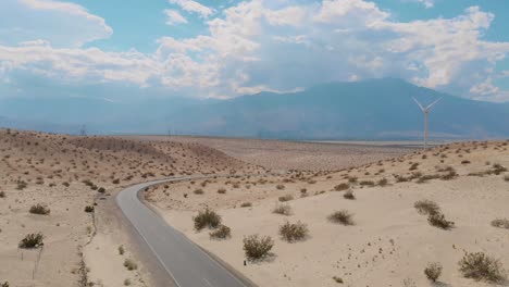 Drone-side-scroll-of-sand-and-brush-covered-hills-near-small-desert-town-and-windmill