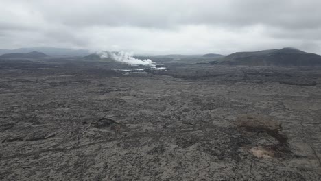 Interminables-Campos-De-Lava-En-Islandia-Con-Humo-Procedente-De-Una-Estación-Geotérmica-Al-Fondo