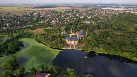 Mogoșoaia-palace-with-adjacent-lake-and-greenery-during-early-morning-light,-outskirts-of-town-in-background,-aerial-view