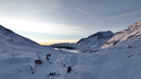 Dawn-on-gorgeous-snow-mountains-with-frozen-lake-and-refuge-house,-Mont-Cenis