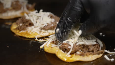 Shredded-cheese-added-to-birria-tacos-cooking-on-flat-top-grill,-black-gloved-hand-sprinkles-grated-quesadilla-cheese-to-sizzling-tacos,-slow-motion-slider-close-up-4K