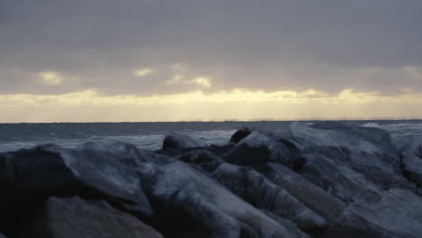 Sanfte-Wellen-Mit-Weißen-Kämmen-Spülen-Sanft-Gegen-Eisige-Felsblöcke,-Während-Die-Sonne-Am-Horizont-Untergeht