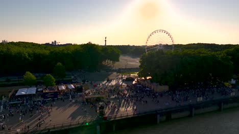 Wine-fair-with-Ferris-wheel-and-crowds-while-sun-sets-behind,-Aerial-pan-left-rising-shot