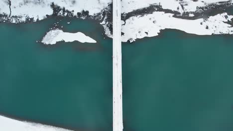 Drone-shot-of-snow-covered-road-over-green-lake-and-snow-covered-landscape