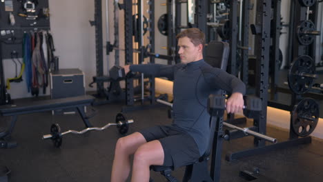 Athletic-blond-man-trains-his-shoulders,-doing-dumbbell-raises-to-the-sides-or-shoulder-fly-exercise-seated-on-bench-in-gym-in-slow-motion