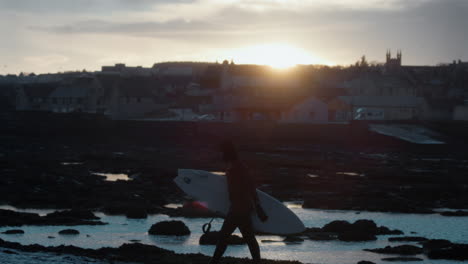 Setting-sun-glistens-above-coastal-town-in-Ireland-as-surfer-in-wetsuit-races-out-of-water-under-storm-clouds