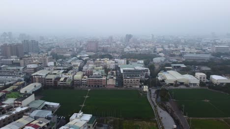 Una-Densa-Zona-Urbana-Con-Edificios-Y-Un-Campo-Verde-En-Un-Día-Nublado,-Atmósfera-Brumosa,-Vista-Aérea