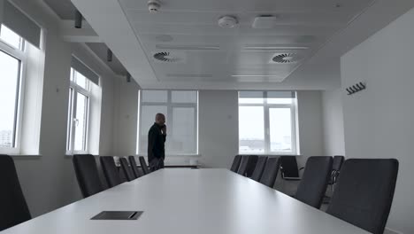 A-man-paces-and-converses-on-his-mobile-phone-in-front-of-an-empty-conference-room-table,-surrounded-by-chairs,-reflecting-the-concept-of-business-communication-and-anticipation