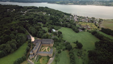 Panoramic-Dolly-Aerial-View-of-Large-Monastery-Building-Surrounded-by-Trees-Uphill-a-Village-by-a-River