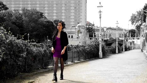 Young-Asian-woman-in-purple-dress,-black-coat-walking-to-the-left-side-of-the-frame-along-the-Wien-River-at-Stadtpark,-a-historical-landmark-featuring-statues-of-famous-Viennese-composers-in-Austria