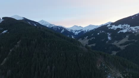 Snow-capped-peaks-tower-over-the-lush-pine-forests-of-Saalbach-Hinterglemm-at-dusk,-aerial-view