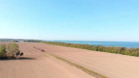 Aerial:-steam-narrow-gauge-railway-in-the-countryside-passing-by-sown-fields-and-the-sea-in-the-background