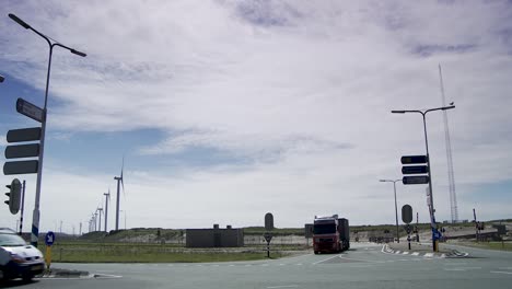 Cars-and-trucks-turning-on-a-roundabout-with-wind-turbines-and-construction-cranes-in-the-background,-daylight
