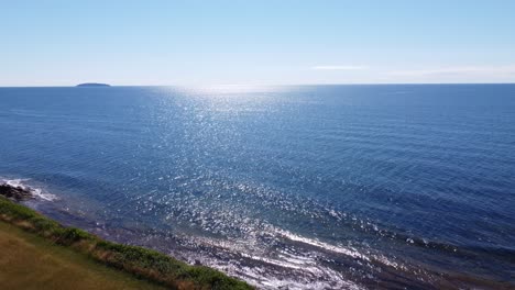 Aerial-drone-flying-over-grass-cliff-revealing-the-ocean-with-clear-blue-sunny-sky-in-the-Maritimes,-Canada
