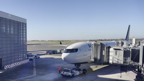 Camión-Pushback-Empujando-El-Avión-De-Air-Canada-Hacia-Atrás-Partiendo-De-La-Puerta-De-La-Terminal-Del-Aeropuerto-Con-Un-Puente-De-Carga-Separado-Y-Operadores-Terrestres-Trabajando-En-El-Asfalto