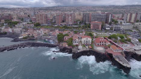 Aerial-shot-of-the-waterfront-of-Catania,-Sicily,-Italy-with-sea-waves-crashing-on-the-volcanic-cliff