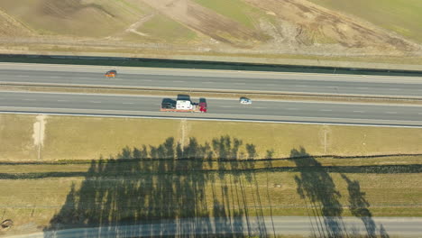 Shadows-stretch-across-a-highway-where-colorful-vehicles-are-spaced-out,-journeying-along-a-road-bordered-by-changing-terrain