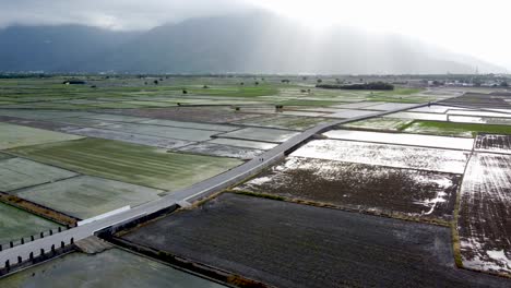 Sunlight-beams-over-wet-paddy-fields-with-mountains-in-the-background,-tranquil-rural-landscape