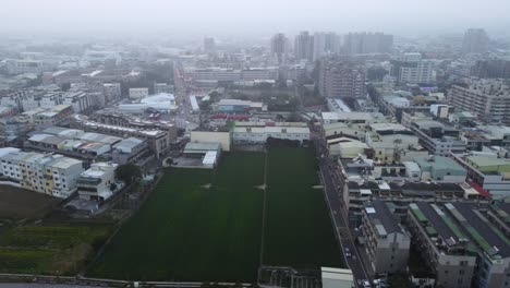 Una-Extensa-Zona-Urbana-Con-Edificios-Y-Un-Campo-Verde-Central-En-Un-Día-Nublado,-Ambiente-Brumoso,-Vista-Aérea