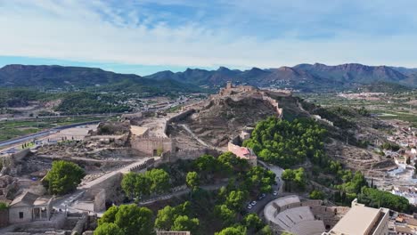 Hochwertige-Aufnahmen-Mit-Einer-Drohne-Der-Burg-Von-Sagunto-Und-Ihren-Mauern-Auf-Einem-Hügel,-Wo-Wir-Auch-Das-Römische-Amphitheater-Mit-Einem-Hintergrund-Aus-Bergen-Und-Einem-Himmel-Mit-Wolken-Sehen,-Valencia,-Spanien