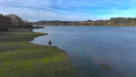 Photographer-With-His-Camera-On-Tripod-Near-Scenic-Lake---Drone-Pullback