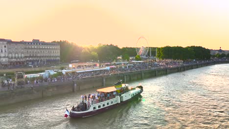 Menschen-Auf-Einem-Ausflugsboot,-Das-Auf-Der-Garonne-Segelt,-Mit-Riesenrad-Und-Sonnenuntergang-Während-Der-Weinmesse,-Luftaufnahme-Nach-Links